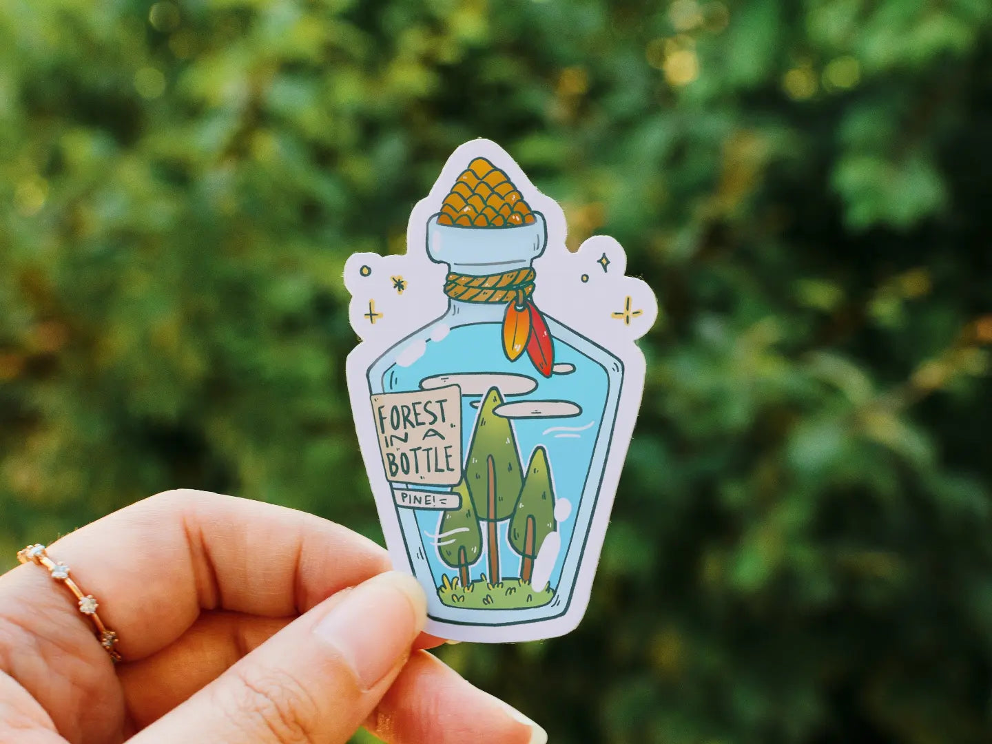 Holding a sticker of a bottle with three trees in it. The label on the bottle reads "Forest in a Bottle".