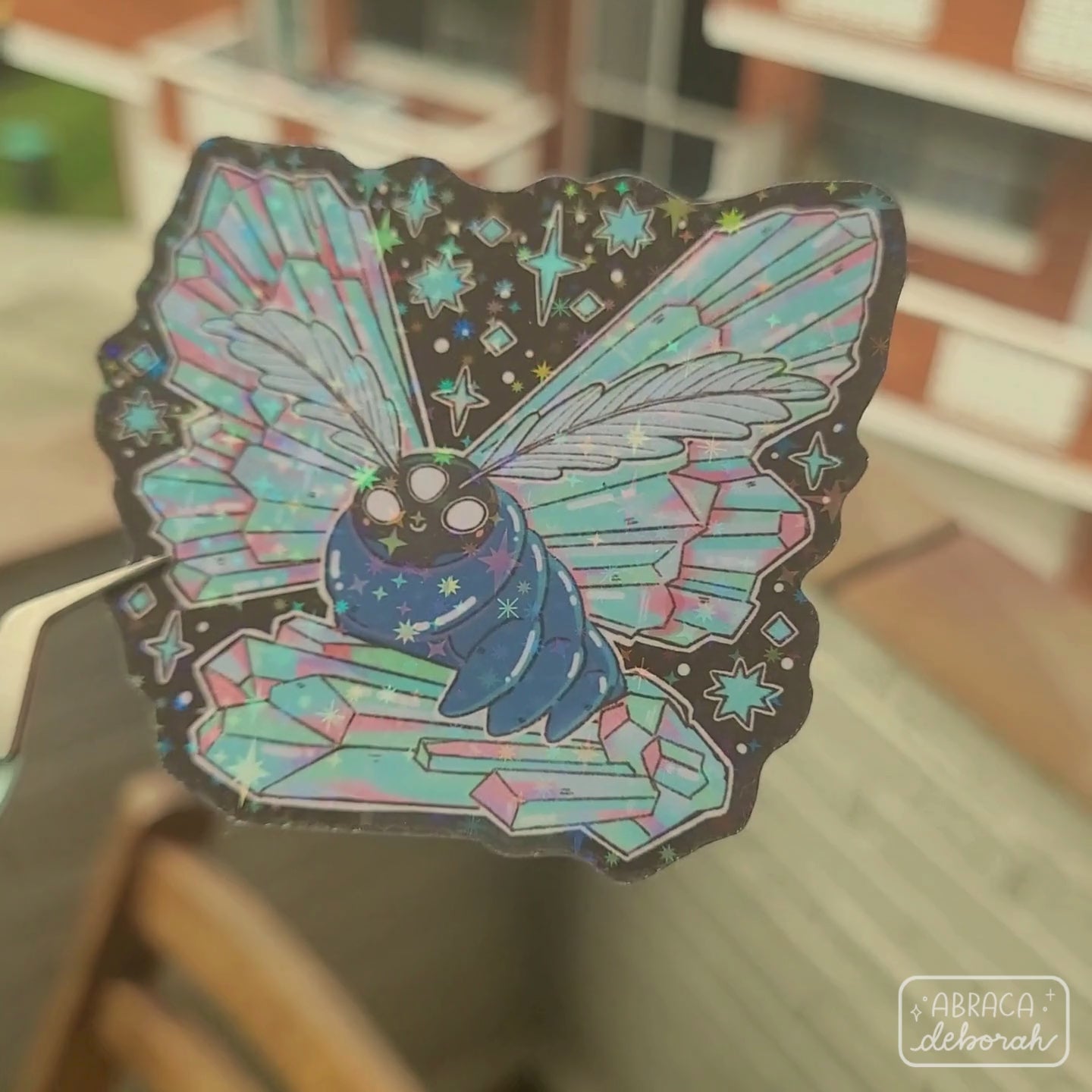 Holographic sticker of a luminous moth with three eyes, featherlike antennae and wings made of iridescent crystals.