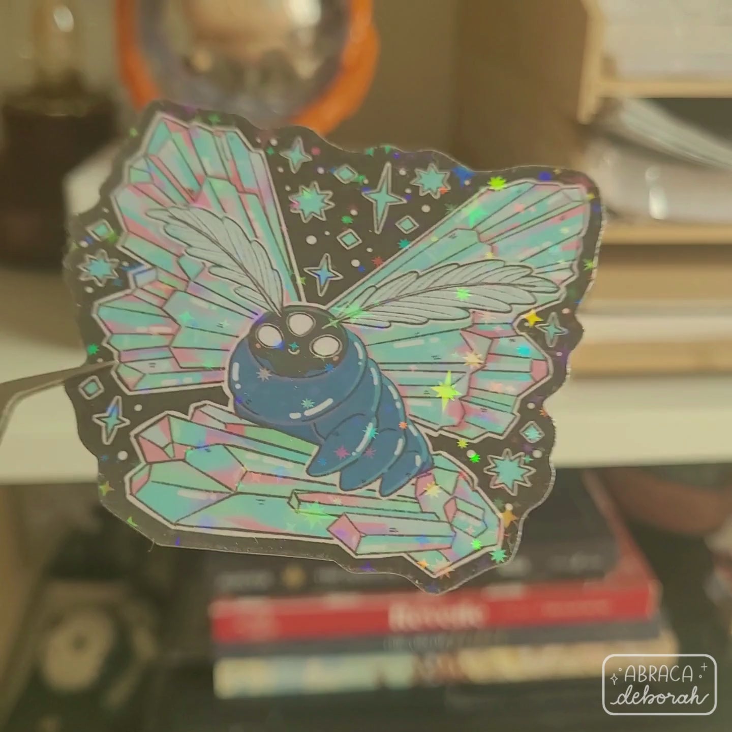Holographic sticker of a luminous moth with three eyes, featherlike antennae and wings made of iridescent crystals.