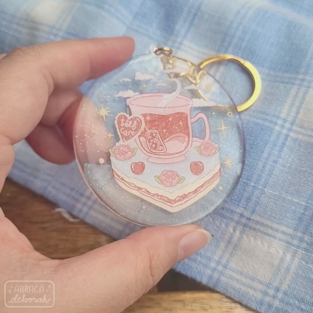 Round acrylic keyring with an illustration of a cup of tea sitting on a slice of cake. The teabag label says "Self Care".