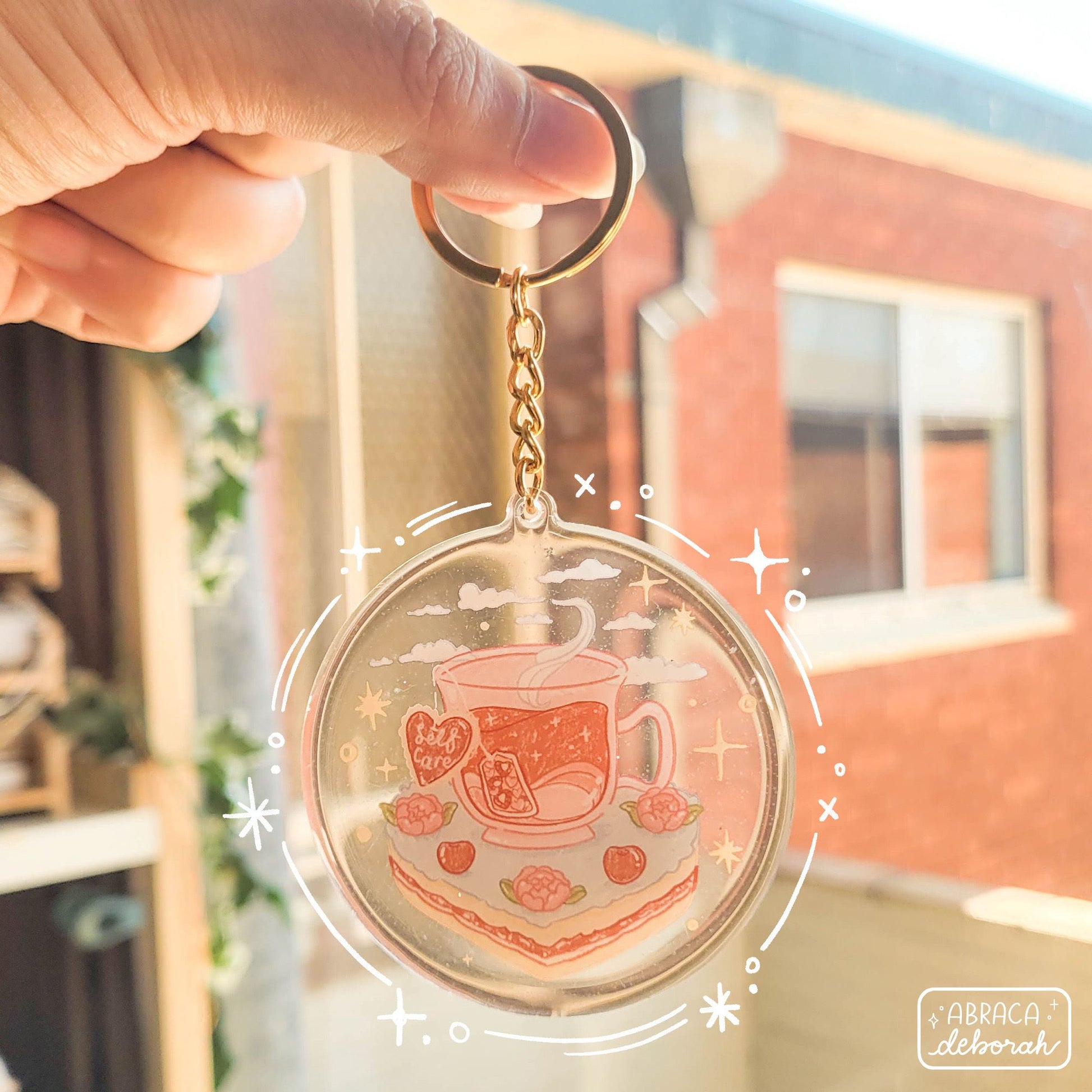 Round acrylic keyring with an illustration of a cup of tea sitting on a slice of cake. The teabag label says "Self Care".