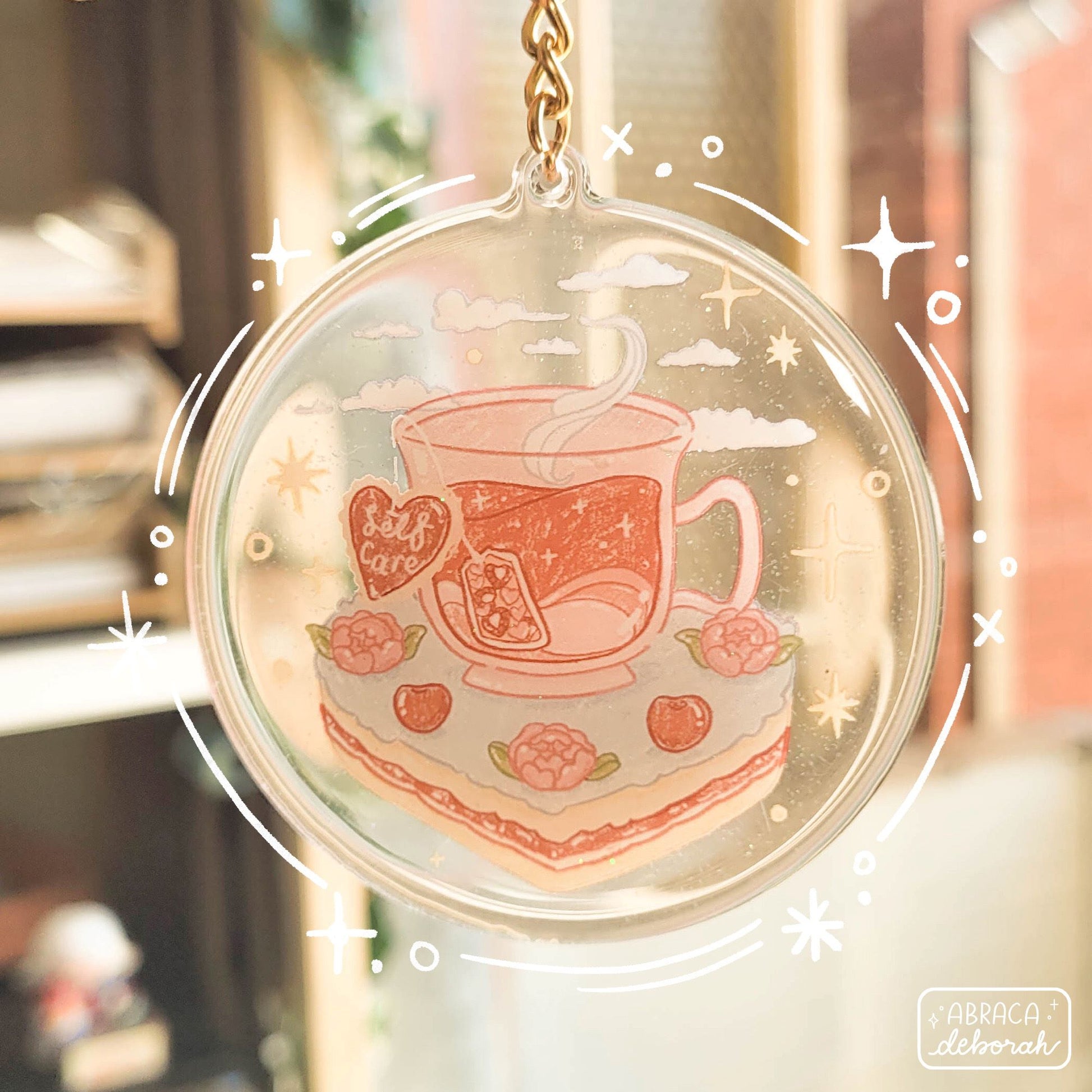 Round acrylic keyring with an illustration of a cup of tea sitting on a slice of cake. The teabag label says "Self Care".