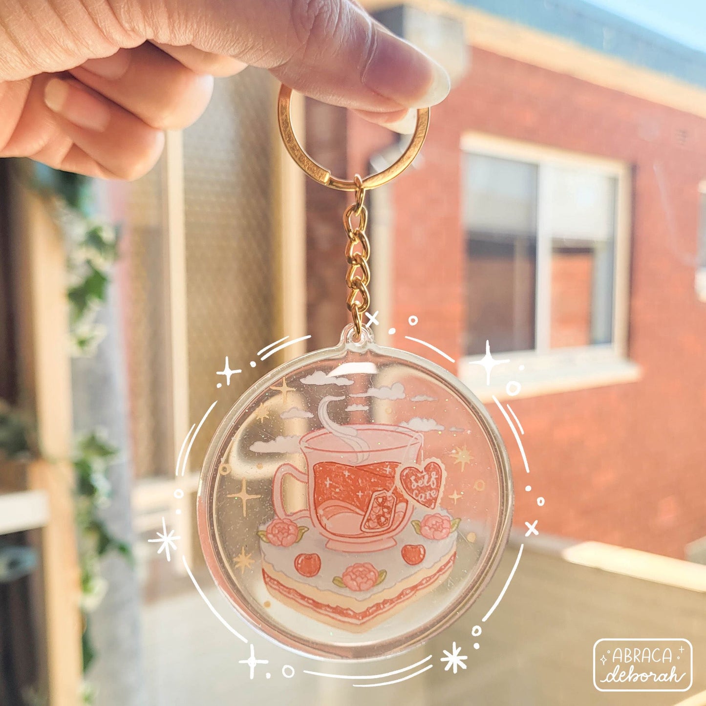 Round acrylic keyring with an illustration of a cup of tea sitting on a slice of cake. The teabag label says "Self Care".