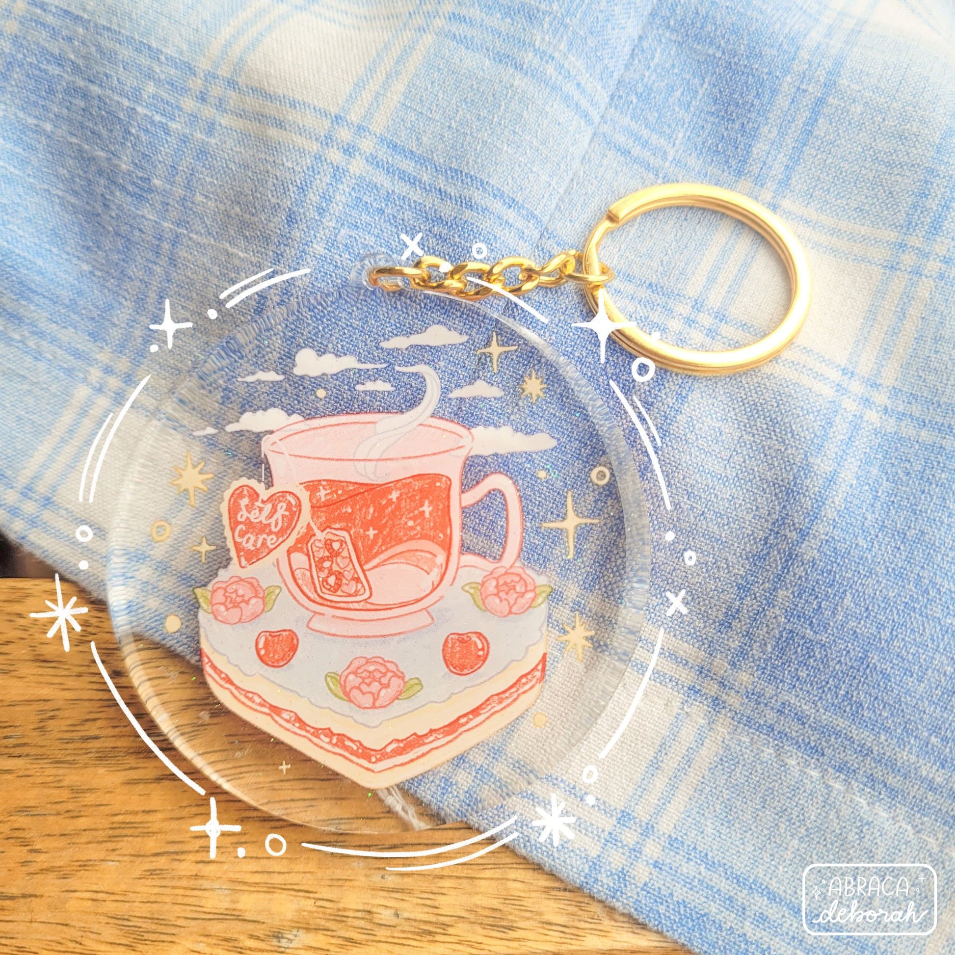 Round acrylic keyring with an illustration of a cup of tea sitting on a slice of cake. The teabag label says "Self Care".