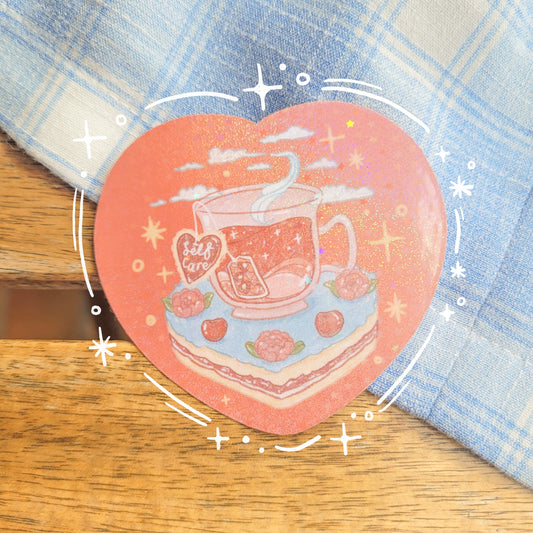 Heart shaped glittery sticker with an illustration of a cup of tea sitting on a slice of cake. The teabag label says "Self Care".