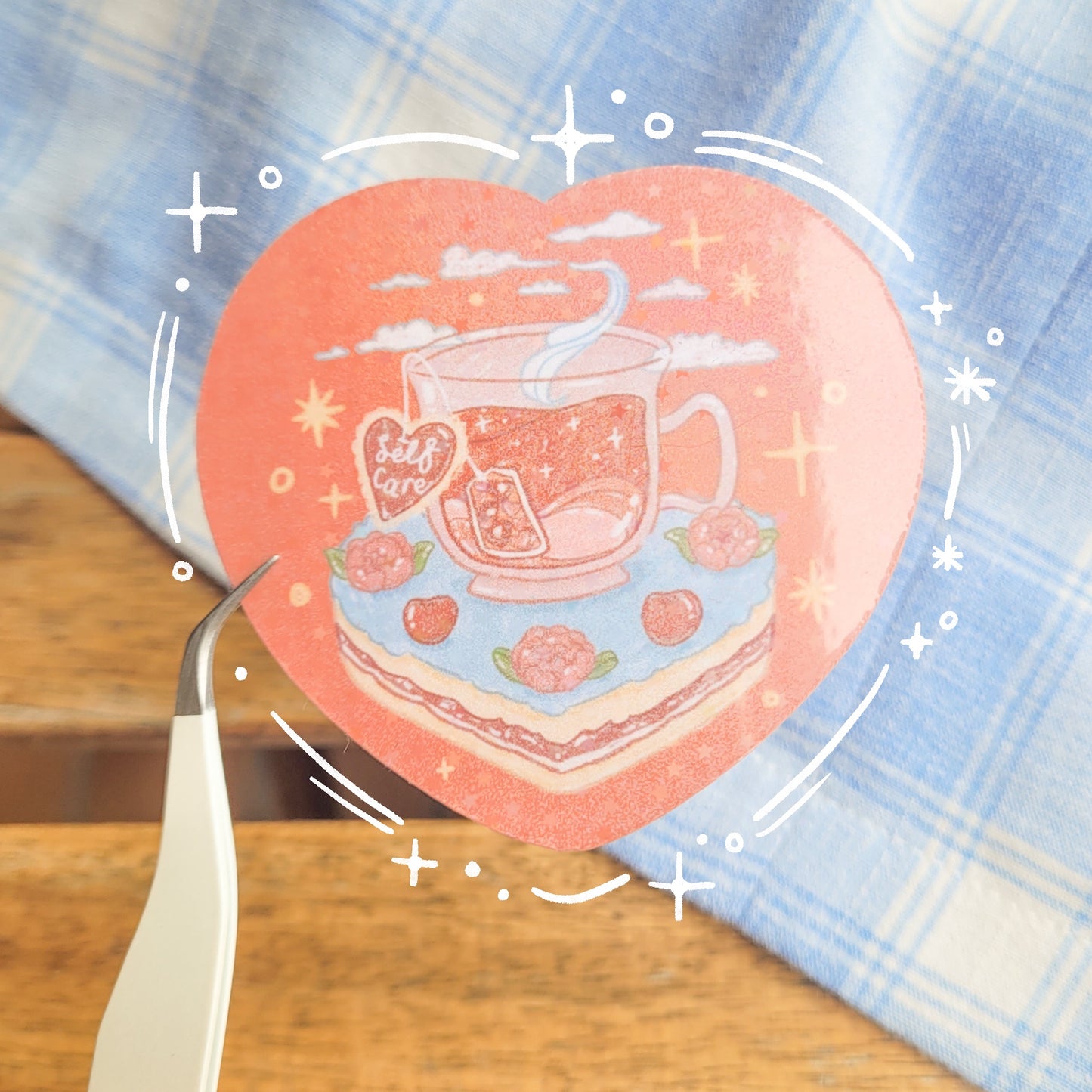 Heart shaped glittery sticker with an illustration of a cup of tea sitting on a slice of cake. The teabag label says "Self Care".