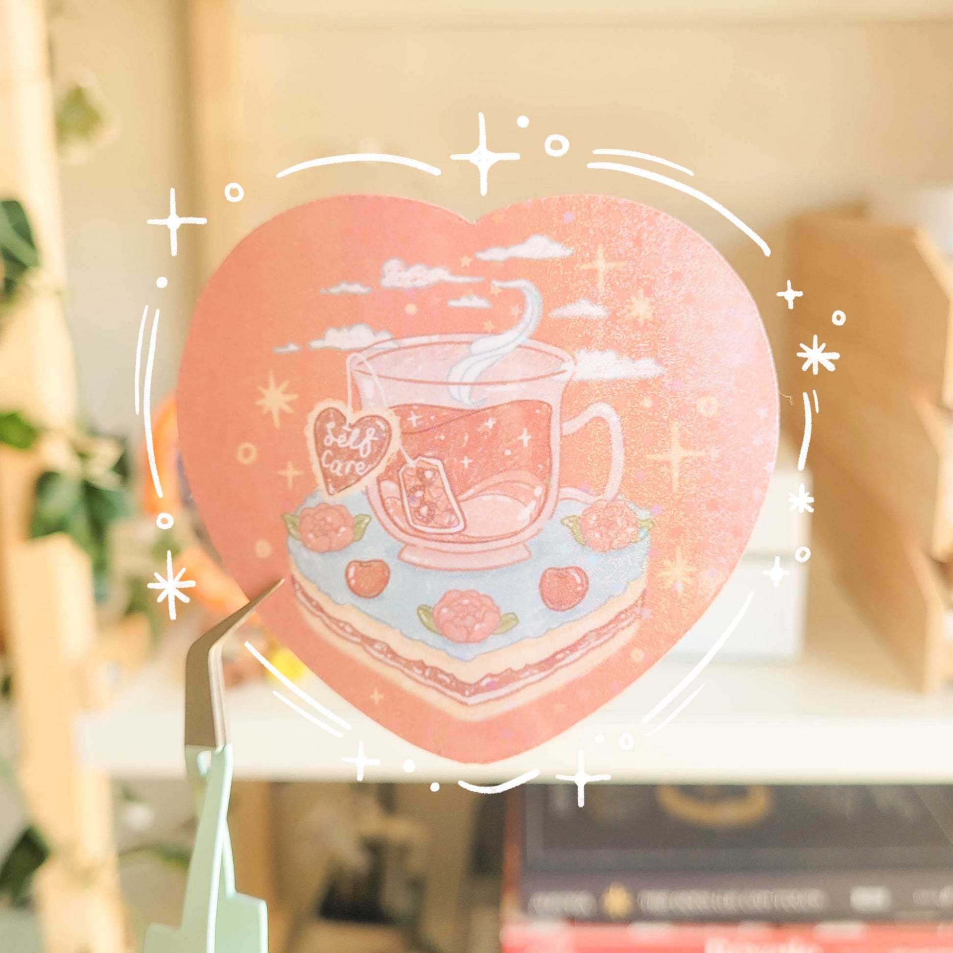 Heart shaped glittery sticker with an illustration of a cup of tea sitting on a slice of cake. The teabag label says "Self Care".