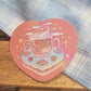 Heart shaped glittery sticker with an illustration of a cup of tea sitting on a slice of cake. The teabag label says "Self Care".